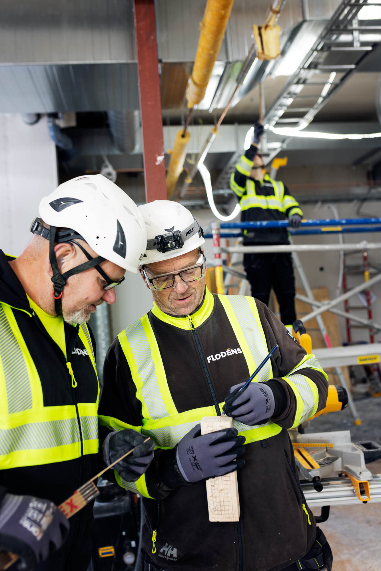Två byggarbetare från Flodéns i reflexkläder och hjälmar granskar en ritning och gör anteckningar på en träbit inne i en industrilokal. I bakgrunden arbetar en tredje person på en byggställning med installation av kablar i taket. Alla bär skyddsutrustning och arbetsmiljön är tydligt en pågående byggarbetsplats.